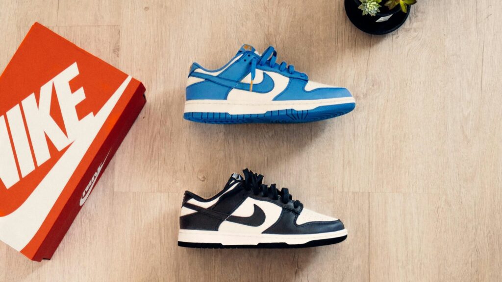 Twee Nike Dunk sneakers in blauw-wit en zwart-wit liggen naast een rode schoenendoos op een houten vloer.