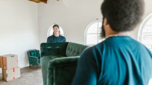 Twee mannen tillen samen een groene fluwelen bank tijdens een verhuizing in een lichte woonkamer met verhuisdozen.